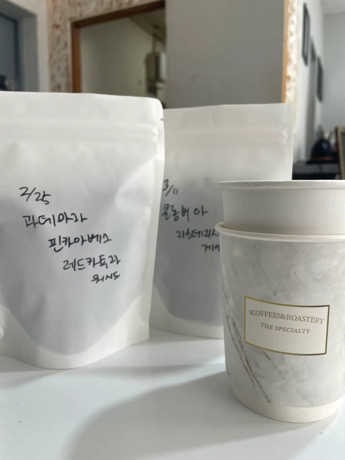 3커피스엔로스터리의 흰 원두 봉투 두 개에 볶은 날짜와 나라, 농장, 품종, 가공 방식이 손글씨로 적혀 있고, 옆에 가게 이름이 찍힌 종이컵이 놓여 있습니다.