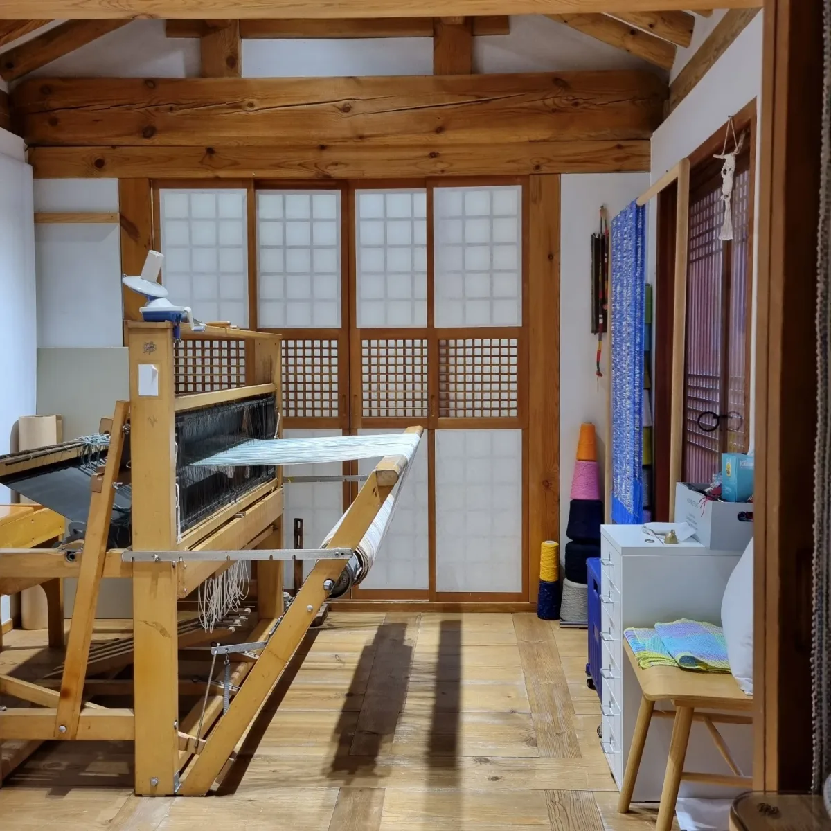 직물놀이공방 한옥 마루에 놓인 나무 수직기와, 뒤편 선반에 색색의 실과 직물이 쌓여 있는 사진입니다.